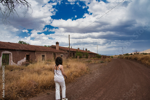 Woman stands on a dirt road, facing abandoned buildings. The sky shows clouds against blue. Her white outfit contrasts the dry landscape. The structures look old and weathered. She gazes at the scene