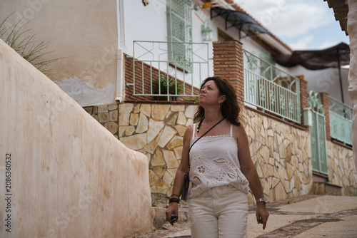 Woman strolls along narrow, sunlit stone alleyway. She gazes upward, smiling softly at sky. Warm tones and rustic walls frame her journey. Perfect for travel, lifestyle, or leisure themes