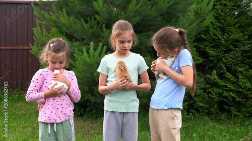 Three happy girls hold and pet little baby rabbits outdoors in garden. Children care pets. Kids play with animals. Agriculture, farming