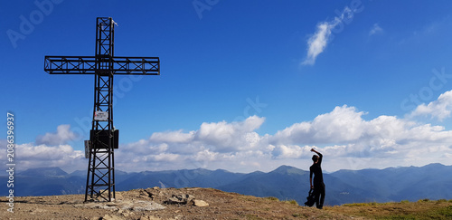 A man stretches near a tall metal cross on a mountain peak surrounded by clear skies and distant hills, creating a serene and reflective atmosphere.