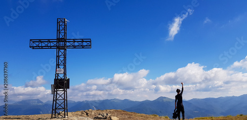 A man raises his arm in joy next to a metal mountain cross, surrounded by breathtaking peaks and a vibrant blue sky with scattered white clouds.