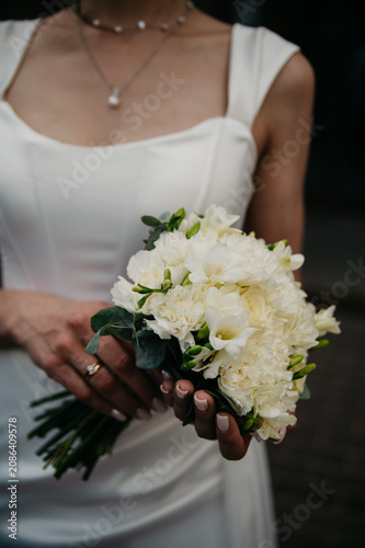 A slender, young and healthy bride in a white fitted wedding dress holds a white bouquet of roses in her hands with a gold wedding ring on her finger and a precious pendant around her neck