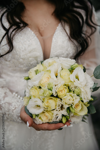 A curvy, young, healthy bride in a white dress embroidered with pearls and a deep neckline with a large bust, holding a bouquet of white roses in her hands