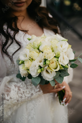 A slender, young, healthy bride in a white dress embroidered with pearls and a deep neckline, with hair falling on her shoulders, holds a bouquet of white roses in her hands