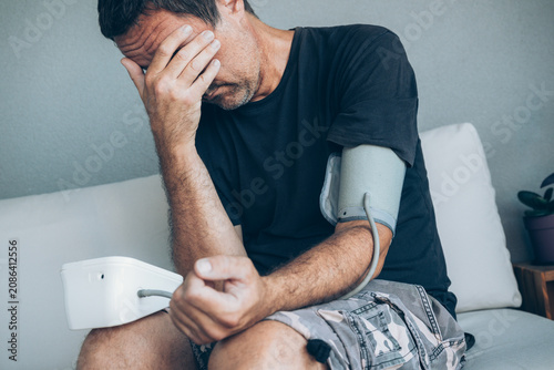 Photos Man controls his blood pressure at home using digital arm cuff while seated in cozy living room, conveying self-care, health monitoring and concern for wellbeing