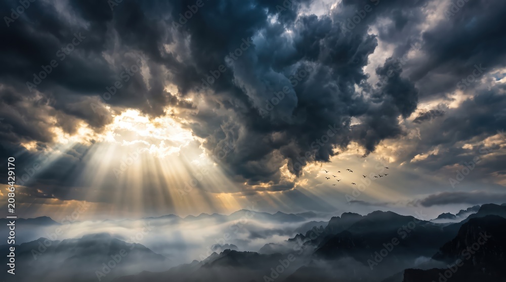 Obraz premium Dramatic mountain landscape with sun rays breaking through storm clouds, fog filled valleys and distant ridgelines with birds in flight, moody atmospheric scene