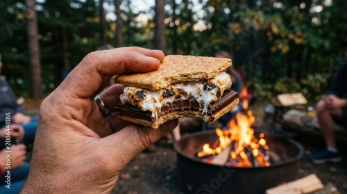 Hand holding a perfectly toasted s'more with melted chocolate and gooey marshmallow by a crackling campfire outdoors.