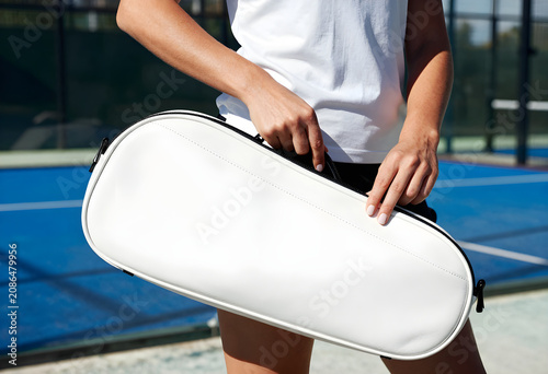 Two hands zipping up a premium white leather padel racket bag against a bright, sunlit sports club wall.