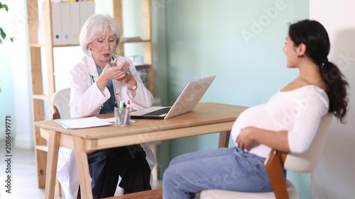Attentive elderly female doctor talking with pregnant patient at medical clinic office table. High quality 4k footage