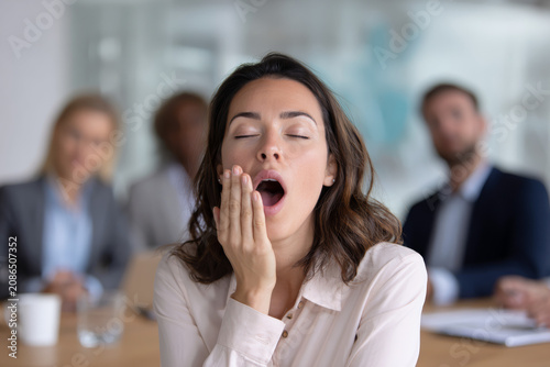 Young woman yawning widely with eyes closed in a business meeting, feeling tired and bored.