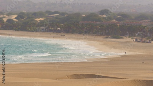 a vast coastal landscape under a hazy, overcast sky. In the foreground, smooth, rolling sand dunes with soft ridges stretch across the scene, transitioning into a wide, sandy beach. 