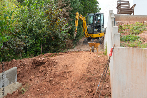 construction workers actively excavating at a residential site using an excavator and wheelbarrows surrounded by concrete blocks and dirt, ground level view, brown and green tones.