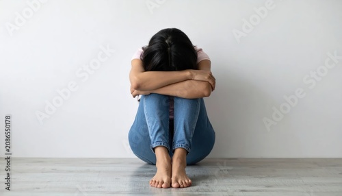 Sad Young Woman Sitting Alone with Head Down Against Plain Wall
