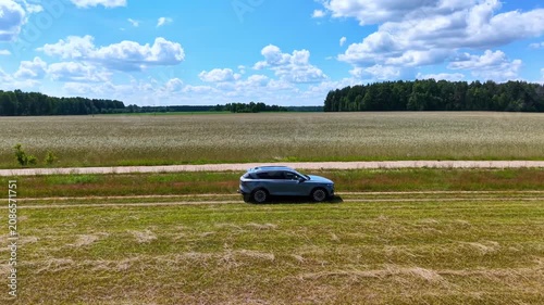Blue SUV parked on grassy roadside next to expansive green field under blue sky, aerial view