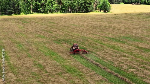 Tractor performing hay cutting on a large grassy field with green trees in the background, aerial view