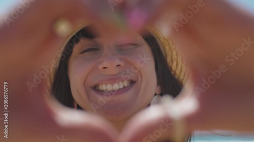 Close-up of a smiling woman on a sunny beach, wearing seashell jewelry. She forms a heart shape with her fingers in the warm sunlight and makes a heart gesture.