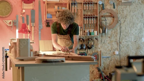 Focused woodworker shaping details on table using plane tool, carefully checks wooden surface, skillful carpenter processing and grounding future chair leg with hand plane, furniture manufacturing.