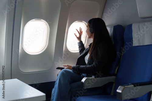 Asian female traveler with headphones waving through window in airplane seat