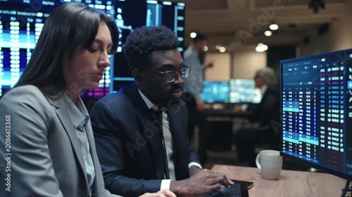 Diverse female and male brokers discussing financial data while looking at computer screen. Business team analyzing stock market charts in dark trading office. Traders working together late at night.