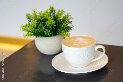 A white cup of hot cappuccino with beautiful latte art on a glossy dark table. Potted green plant in the background, cozy cafe atmosphere, copy space.