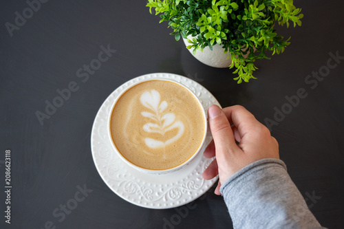 High-angle shot of a person in a grey sweater holding a  cup with latte art, next to a potted green plant on a black background. Coffeebreak concept