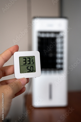 Foto Hand holding a digital room thermometer showing high temperature of 33°C against a blurry portable air cooler background