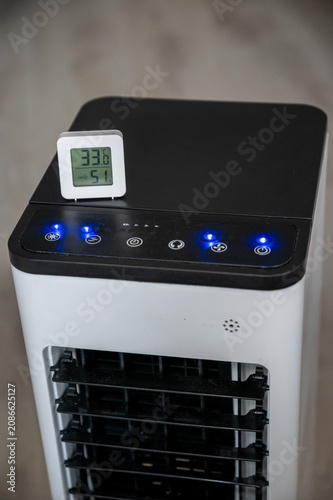 A digital room thermometer displaying a high temperature of 33°C, set against the blurred background of a portable air cooler. Summer heat and the concept of climate control