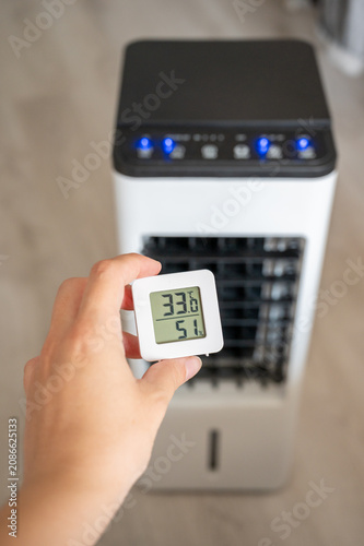 Hand holding a digital room thermometer showing high temperature of 33°C against a blurry portable air cooler background. Summer heat and climate control concept
