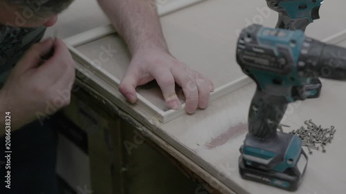 Close-up of a worker assembling a wood frame window on a workshop bench using a cordless drill and screws. DIY carpentry, woodworking, manufacturing, and construction concept