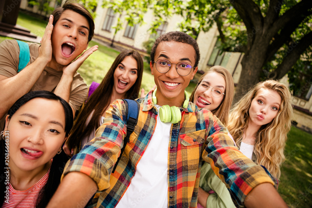 Fototapeta premium Group of happy diverse college friends taking a selfie on a leafy campus, smiling students with backpacks and headphones enjoying outdoor time together