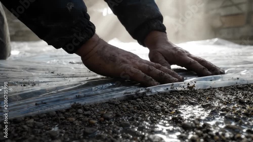 Construction worker smoothing a protective plastic sheet over wet gravel on a building site. A close up shot capturing hands preparing ground insulation, representing hard labor and maintenance.