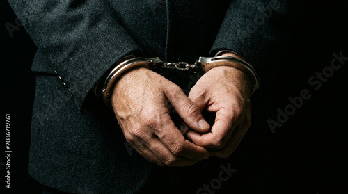 Handcuffed businessman in dark suit, arrested corporate criminal in metal handcuffs, white collar crime and financial fraud concept
