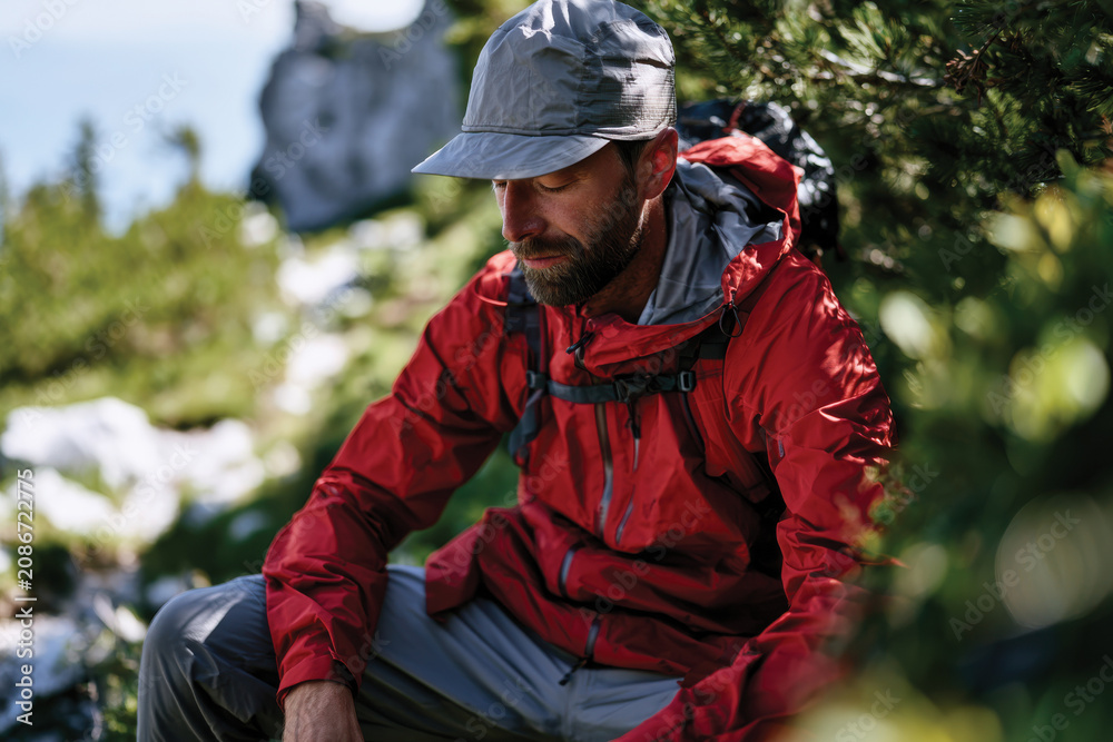 Fototapeta premium In a serene nature setting, a man sits and rests under a tree while wearing a bright red jacket, embodying the spirit of exploration and the beauty of solitude in nature.