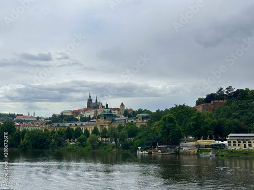 Prague Castle overlooks the Vltava River. Historic waterfront buildings sit under a cloudy blue sky.
