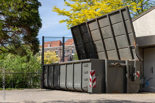 Large industrial waste container with an open lid standing outdoors near a building and green trees. Urban service scene showing waste management equipment and recycling infrastructure .
