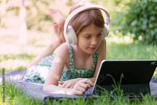 A young girl lying on a blanket in a park, using a tablet with headphones, concept of digital learning outdoors.
