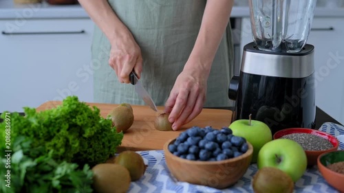 Woman slicing fresh kiwi on wooden cutting board beside blender, blueberries, apples and leafy greens for green smoothie. Woman prepares nutritious ingredients supporting healthy lifestyle 