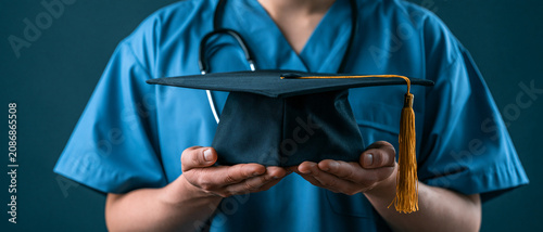  Close-up of an unrecognizable medical student in blue scrubs with a stethoscope holding a black graduation cap. Medical education and healthcare career concept.