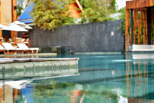 Luxurious tropical resort swimming pool with lounge chairs and umbrellas surrounded by beautiful green trees and architecture.