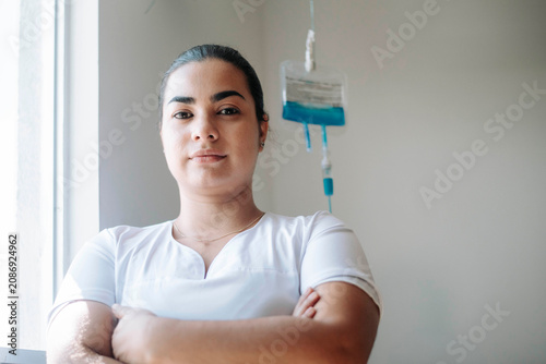 Confident female nurse standing beside an intravenous IV drip with folded arms in a modern clinical environment.