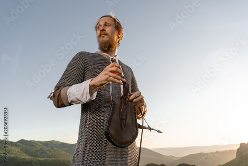 Foto A bearded man in chainmail and tunic stands on a hilltop, gazing toward distant mountains at sunset with a leather pouch slung over his shoulder