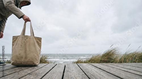 Person holding a blank reusable canvas tote bag on a weathered wooden deck by the windy cloudy coast
