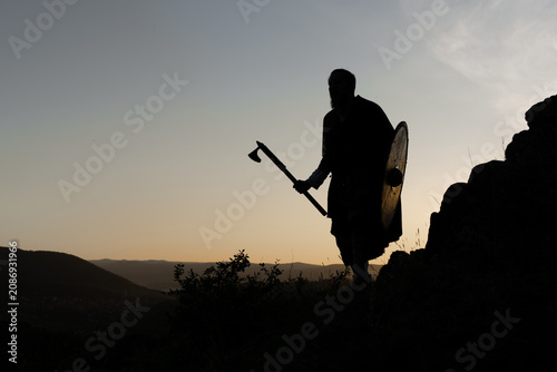 Photos Silhouetted Viking stands atop a rocky outcrop at sunset, clutching an axe and shield against a vast sky