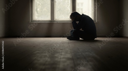 Silhouette of man sitting on floor in dark empty room. Person with head in hands by window. Concept of depression and loneliness