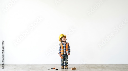 Young child in construction worker outfit standing in an empty room with toy tools.