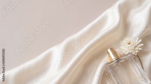Clear glass perfume bottle and dry white flower on elegant cream silk fabric with soft flowing folds