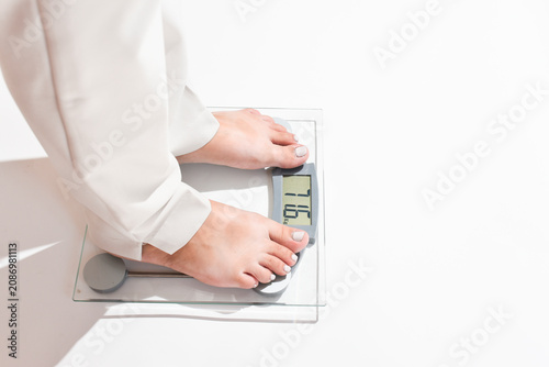 Woman’s legs in white pants step onto a bathroom scale on a clean white background. Close-up of feet with a light gel pedicure standing on digital scales. Weight control, weight loss journey