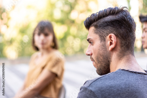 Serious young man in profile during a tense outdoor conversation. Blurred friends sit nearby at a patio table with soft bokeh. Relationship conflict concept
