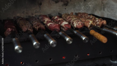 A close-up cinematic shot of raw pork and chicken meat skewers grilling over hot charcoals inside a barbecue, with handles in focus and heat glowing underneath