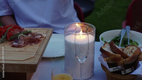 A close-up cinematic shot of a romantic candlelight dinner setup outdoors, featuring a burning candle in a glass holder, a hand holding a champagne flute, and gourmet appetizers.